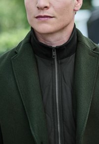 Mörkgrön ullblandad kappa över en svart zip-up-jacka med kviltad textur, med ståkrage och silverdragkedja.