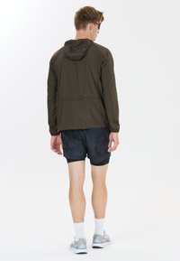 Mann trägt dunkle Kapuzenjacke, schwarze Sportshorts, weiße Socken und graue Laufschuhe, steht mit dem Rücken zur Kamera vor einfarbigem Hintergrund.