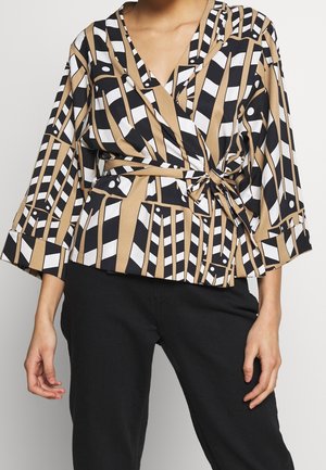 Woman wearing a beige, black, and white geometric print wrap blouse with three-quarter sleeves and black pants.
