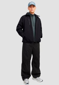 Svart zip-up jakke over en grønn genser, sammen med løse svarte bukser. Modellen har på seg en lyseblå caps og sølvfargede sneakers.