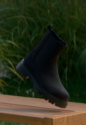Black ankle boot with thick ridged sole hovering above a wooden bench, with green plants in the blurred background.