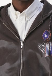 Sudadera con cremallera negra y patrón floral blanco, con capucha, superpuesta a una camisa blanca abotonada, que presenta una cremallera plateada y un tejido texturizado.