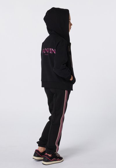 Sweatshirt noir avec capuche, présentant un logo rose au dos. Pantalon de jogging noir avec une bande en zigzag rose. Chaussures noires avec des accents roses.