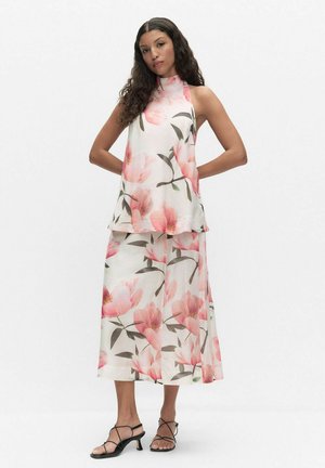 Kobieta o długich, kręconych włosach ubrana w dopasowany, bezrękawnik z wysokim kołnierzem oraz szerokie spodnie z dużym różowym kwiatowym wzorem, stoi z rękami za plecami.