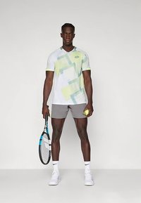 Λευκό και ανοιχτό πράσινο αθλητικό πουκάμισο με σύντομα μανίκια, συνδυασμένο με γκρι σορτς. Κρατά ρακέτα τένις και μπάλα.