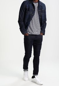 Chaqueta de mezclilla oscura con dos bolsillos en el pecho sobre una camiseta de rayas. Jeans oscuros de corte ajustado y zapatillas blancas con detalles en negro.