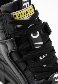 Černá kožená bota s tlustými černými tkaničkami, žlutou značkou "BUFFALO" na jazyku a výrazným bílým písmem na chrániči tkaniček.