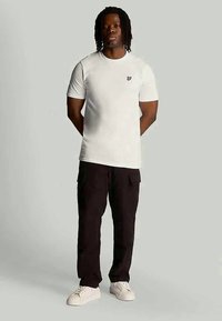 Hvid kortærmet t-shirt med et lille logo på brystet, parret med sorte cargo-bukser og hvide sneakers, stående foran en grå baggrund.