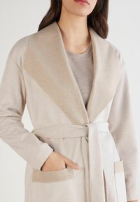 Beżowy długi kardigan z szerokim kołnierzem, wiązaniem w talii i przednimi kieszeniami. Miękka tekstura z kontrastującym kołnierzem i akcentami kieszeni.