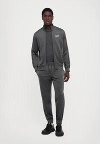 Chándal deportivo gris hecho de tejido suave, con chaqueta con cremallera y cuello, pantalones a juego y zapatillas negras. Logo en el pecho.