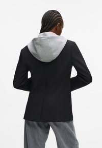 Čierny upravený blejzer so šedou kapucňou. Blejzer má slim fit, tvarované plecia a hladkú textúru, čo ho robí ideálnym na rôzne štýlové kombinácie.