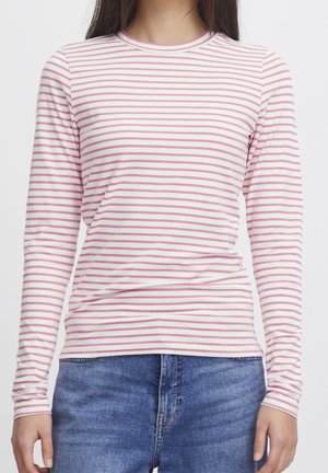 Kobieta w białej bluzce z długim rękawem ozdobionej cienkimi różowymi poziomymi paskami oraz w niebieskich dżinsach, pokazana od szyi do bioder.