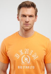 Oranžinė medvilninė marškinė su balta spausdinta dizainu, kuriame yra tekstas "DENIM" ir "BIG STAR LTD" apskritimo raštu su laurų akcentais.