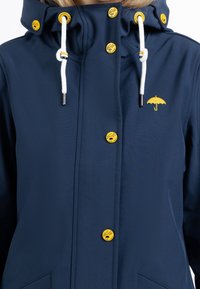 Veste imperméable marine avec col montant, boutons jaunes, cordon blanc, et petit logo parapluie jaune sur la poitrine.