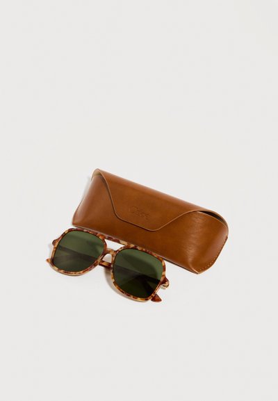 Τετράγωνα γυαλιά ηλίου με σκελετό χελώνας και σκούρους πράσινους φακούς δίπλα σε καφέ δερμάτινη θήκη με ανάγλυφο όνομα μάρκας.