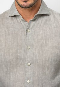 Camicia di lino grigio chiaro con colletto standard, sette bottoni sul davanti e una texture sottile con lievi variazioni di colore.