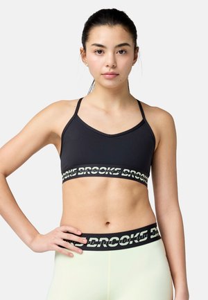 Joven mujer con un sujetador deportivo negro y leggings de color claro con el logo "Brooks", de pie con la mano en la cadera, mirando hacia adelante sobre un fondo blanco.