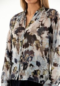 Blusa translúcida con estampado floral en tonos de azul, marrón y verde. Cuenta con un cierre de botones en la parte delantera y mangas largas con puños.