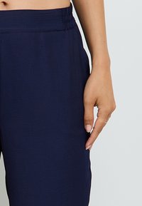 Pantalons bleu marine à taille élastique en tissu doux, avec une coupe ample et des poches latérales, avec une main reposant sur la ceinture.