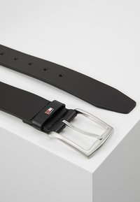 Ceinture en cuir noir avec une boucle en métal argenté rectangulaire, présentant cinq trous pour l'ajustement et un petit logo accent près de la boucle.