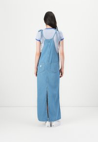 Maxi abito in denim con spalline regolabili, due tasche posteriori e una fessura frontale. Colore blu chiaro, texture liscia.