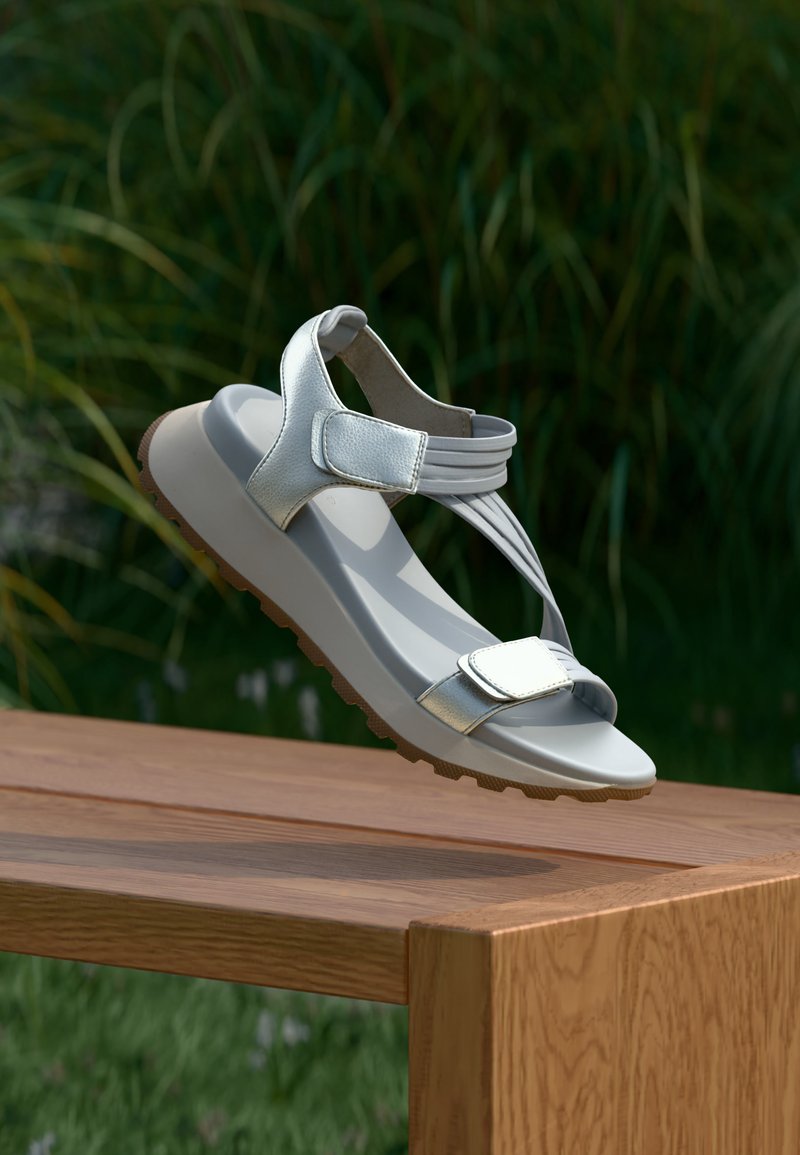 Sandalia plateada con tiras de velcro y suela de goma texturizada suspendida sobre un banco de madera con un fondo de follaje verde.