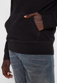 Osoba w czarnej bluzie z kapturem, z jedną ręką w przedniej kieszeni i w zblakniętych niebieskich dżinsach, stojąca na tle jasnego, jednorodnego tła.