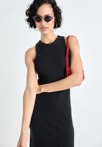 Μαύρο αμάνικο φόρεμα με εφαρμοστή ραβδωτή υφή. Το μοντέλο φοράει σκούρα γυαλιά ηλίου και κρατάει μια μικρή κόκκινη τσάντα με λουράκι στον ώμο.