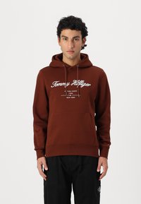 Brun genser med hette og snor, frontlomme og hvit "Tommy Hilfiger" logo tekst. Glatt tekstur med ribbestrikkede mansjetter og nederkant.