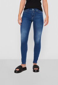 Jeans ajustados en denim azul oscuro con una textura suave, cinco bolsillos y una cintura de altura media, combinados con sandalias deslizantes tejidas en negro.