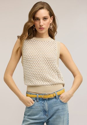 Vrouw met golvend lichtbruin haar, gekleed in een beige mouwloze gebreide top, gele riem, blauwe spijkerbroek en oorbellen in de vorm van bladeren, met handen in de zakken.