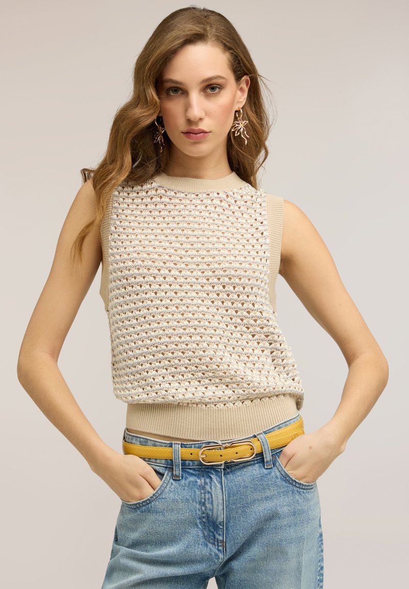 Vrouw met golvend lichtbruin haar, gekleed in een beige mouwloze gebreide top, gele riem, blauwe spijkerbroek en oorbellen in de vorm van bladeren, met handen in de zakken.