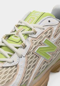 Κοντινό πλάνο από ένα μπεζ και πράσινο αθλητικό παπούτσι New Balance με λευκά κορδόνια, λεπτομέρειες από δίχτυ και χοντρή σόλα.