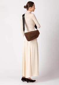 Braune Wildleder-Umhängetasche mit geschwungener Form, verstellbarem schwarzem Riemen und silberfarbenen Details, getragen über einem hellfarbenen Kleid.