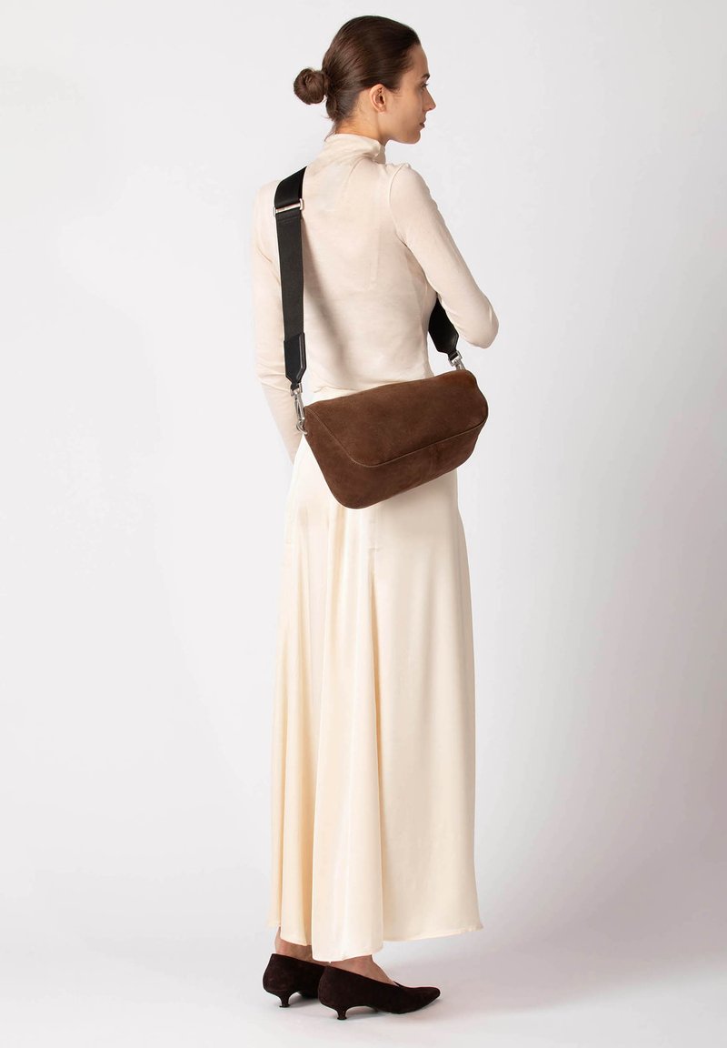 Braune Wildleder-Umhängetasche mit geschwungener Form, verstellbarem schwarzem Riemen und silberfarbenen Details, getragen über einem hellfarbenen Kleid.
