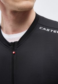 Nærbilde av en person som har på seg en svart sportsjakke med glidelås og "CASTELLI" på skulderen, med en hvit mesh-undertrøye synlig ved kragen.