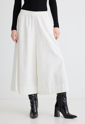 Persona con pantalones anchos blancos recortados con cintura elástica y botas negras de tacón sobre un fondo claro y liso.