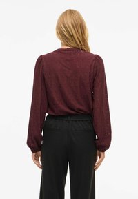 Bordeauxrød bluse med lange ærmer og tekstureret diamantmønster, med puffede skuldre og elastisk talje, kombineret med sorte bukser med høj talje.