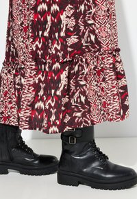 Botas de cuero negro con cordones y hebillas, llevadas debajo de una larga falda de estampado rojo y blanco, sobre un fondo blanco.