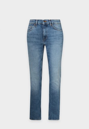 Ung mand stående iført en simpel hvid t-shirt, blå jeans og beige sneakers mod en hvid baggrund.