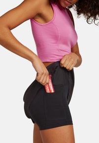 Svarta träningsshorts med en ficka som håller en rosa energibar. Modellen bär en ljusrosa åtsittande linne med texturerade detaljer.