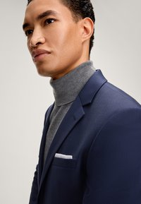 Dunkelblauer, strukturierter Blazer mit Reverskragen, kombiniert mit einem grauen Rollkragenpullover. Ein weißes Einstecktuch hebt die Brusttasche des Blazers hervor.