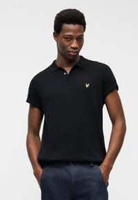 Άνδρας που φοράει μαύρο πόλο με κοντά μανίκια και μικρό χρυσό λογότυπο αετού, μαζεμένο μέσα σε σκούρο μπλε τζιν, με τα χέρια στις τσέπες.