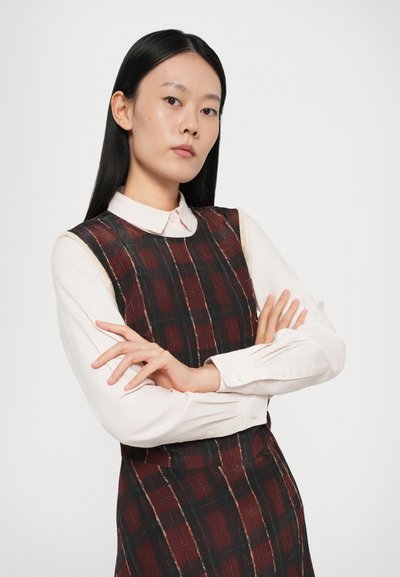 Abito a scacchi nero e bordeaux con design senza maniche sovrapposto a una camicia bianca con colletto. Texture liscia con dettagli di cucitura visibili.