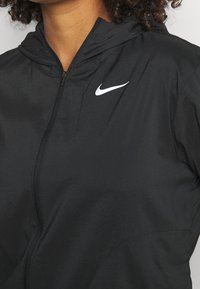 Mustat kevyet vetoketjulliset takki hupulla, jossa on teksturoitu kangas ja valkoinen Nike-logo vasemmassa rinnassa.