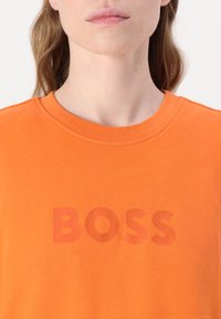 Oranžové bavlnené tričko s okrúhlym výstrihom a slovom "BOSS" potlačeným o niečo tmavším odtieňom na prednej strane. Hladká textura.