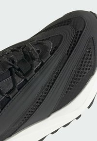 Zapatilla negra con diseño de malla, texturas en capas y una suela de goma prominente con surcos distintos. Sistema de cordones integrado en la parte superior.