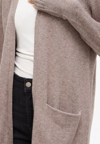 Beige cardigan, mjuk stickad textur, öppen framdesign, långa ärmar med ribbade manschetter, två framfickor. Bärs över en vit topp.