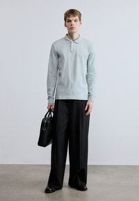 Μακρυμάνικο πόλο σε ανοιχτό γαλάζιο χρώμα με μικρό λογότυπο, συνδυασμένο με μαύρο παντελόνι φαρδιάς γραμμής και μαύρη ορθογώνια τσάντα. Ομαλή υφή υφάσματος.