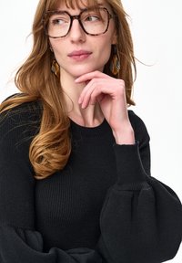 Kobieta o długich, jasnobrązowych włosach, nosząca okulary w oprawkach w panterkę, duże wzorzyste kolczyki oraz czarny prążkowany sweter z bufkami na rękawach, ręka przy brodzie.